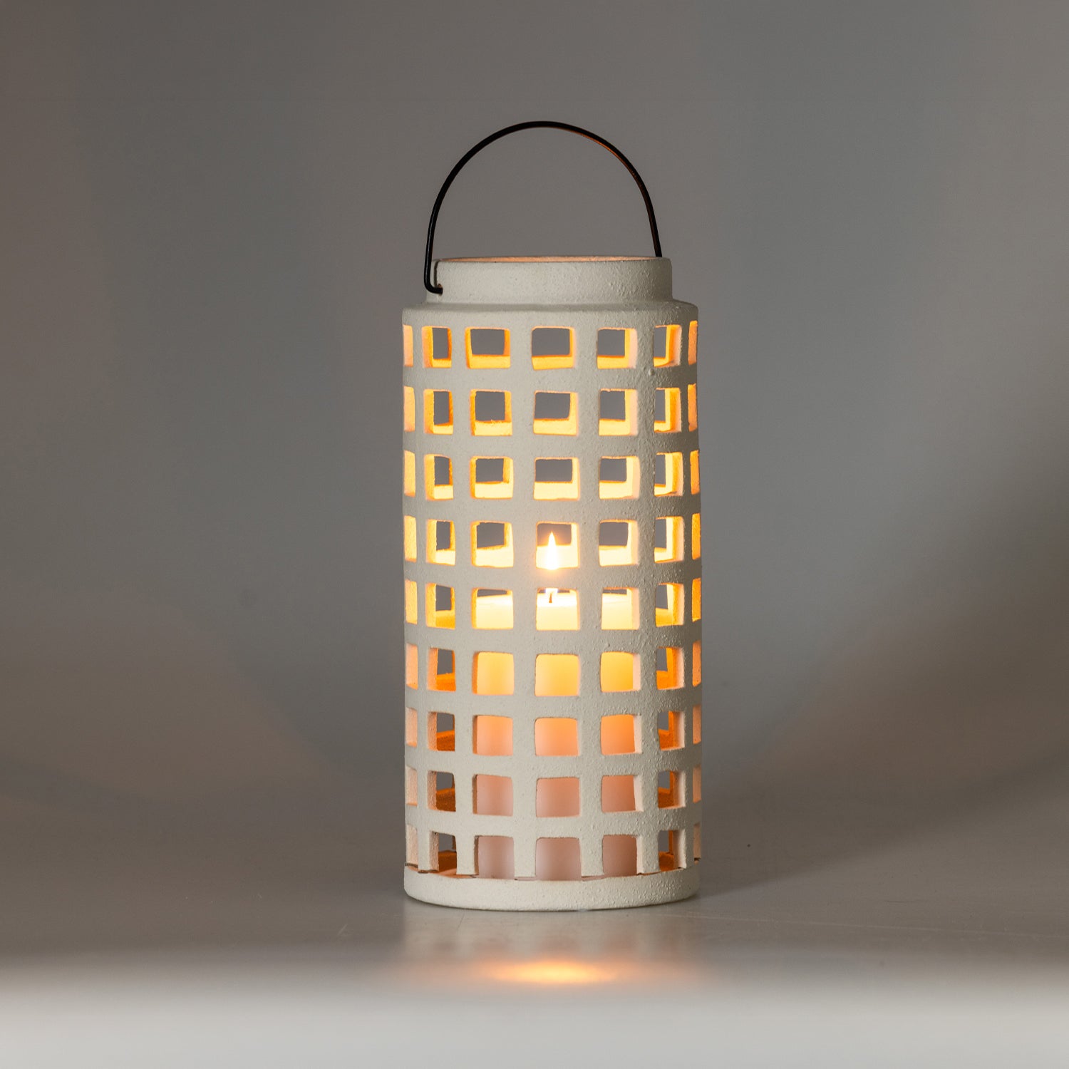 Illuminated white ceramic lantern with a lit candle casting a warm glow through the square cut-out pattern against a soft neutral background.