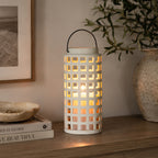White ceramic lantern with a glowing candle displayed on a wooden console beside stacked books, a decorative tray and a large ceramic vase with olive branches.