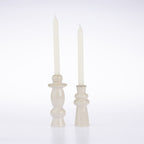 Two cream-colored glass candlestick holders with tall taper candles, set against a seamless white studio background.