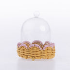 A clear glass cloche dome with a small round knob on top, resting on a round woven wicker base with a scalloped edge of light purple ribbon, photographed against a clean white background.