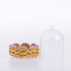 A clear glass cloche dome with a round knob lid stands next to a small woven wicker basket holding several pink and lavender spiral hair extensions on a white background.