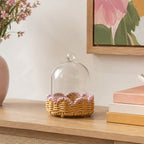 A clear glass cloche dome with a small knob top sits on a woven wicker base decorated with pink ruffled trim, displayed on a wooden console table in a styled interior setting.