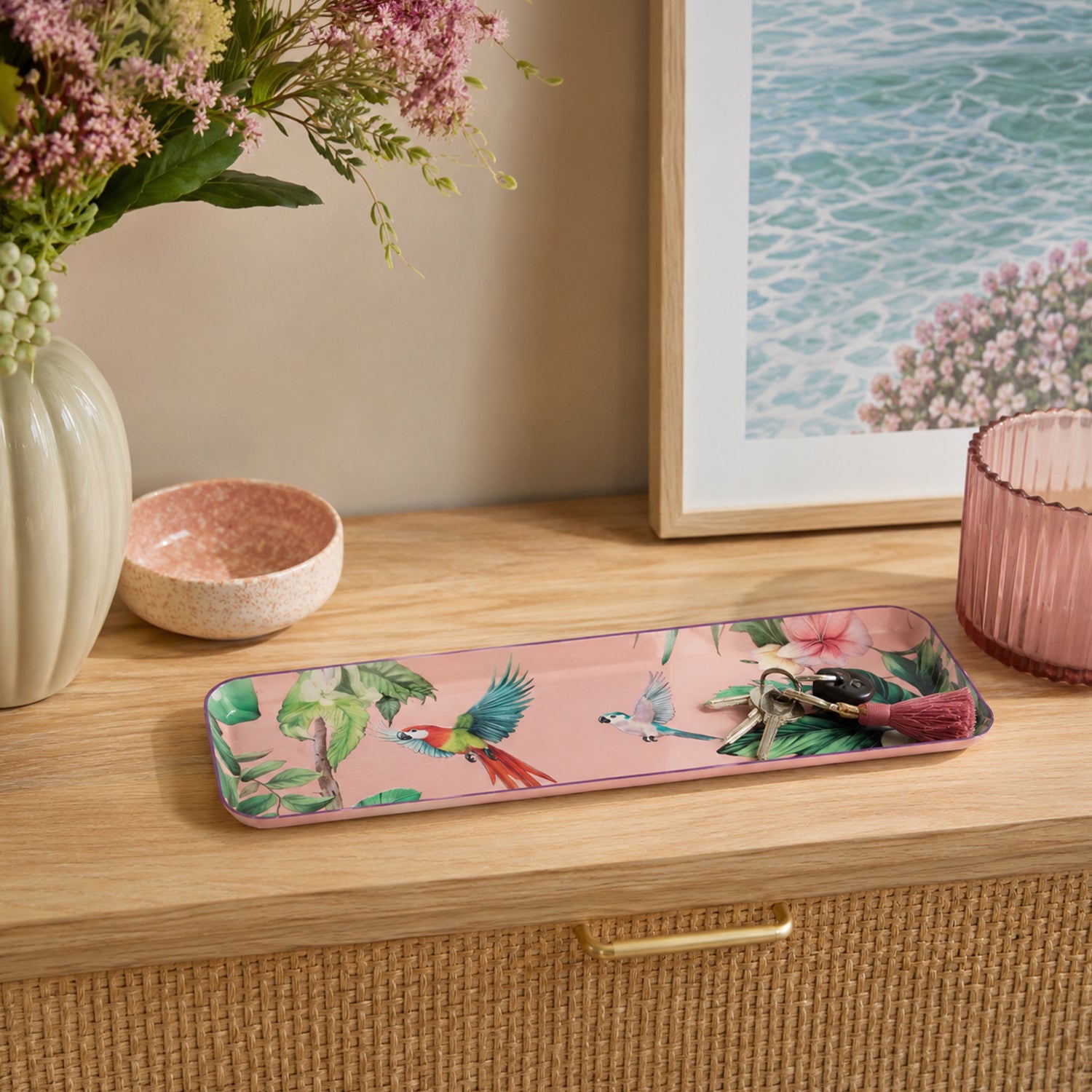A rectangular pink tray with a tropical bird and floral print, styled on a light wood dresser alongside a pink bowl, a vase of flowers, and a framed picture.