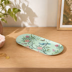 A long, oval ceramic tray with a light mint green background, decorated with a delicate floral and bird pattern in pink, blue, and green, resting on a light wood tabletop.