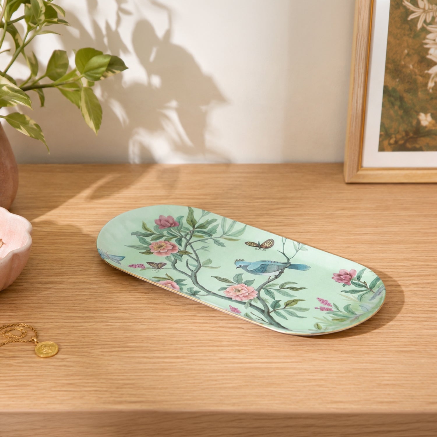 A long, oval ceramic tray with a light mint green background, decorated with a delicate floral and bird pattern in pink, blue, and green, resting on a light wood tabletop.
