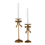 A pair of ornate gold-toned candlesticks with bow details, each holding a white taper candle, shown against a plain white background.