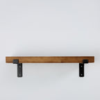 A rustic wooden floating shelf with two black metal brackets, shown against a plain light gray wall.