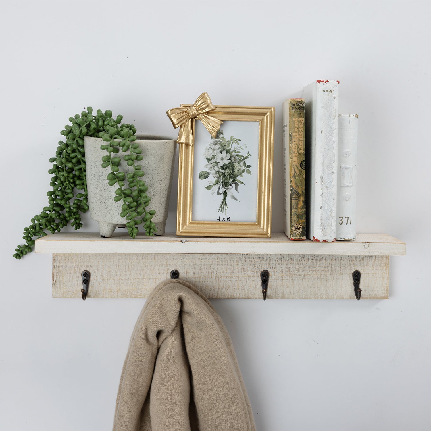A rustic white-painted wooden wall shelf mounted on a white wall, holding a potted trailing green plant, a small gold picture frame with a bow, books, and a beige towel hanging from black hooks.
