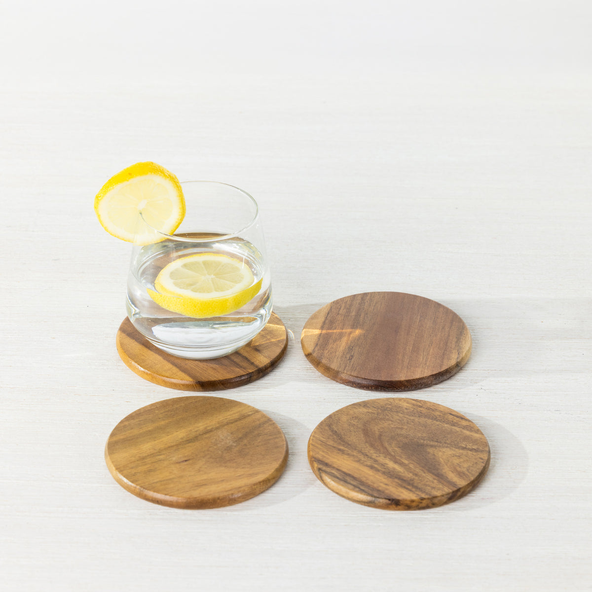 Gather 4 Piece Acacia Coaster Set featuring four round wooden coasters with natural grain patterns, styled with a glass of lemon water.