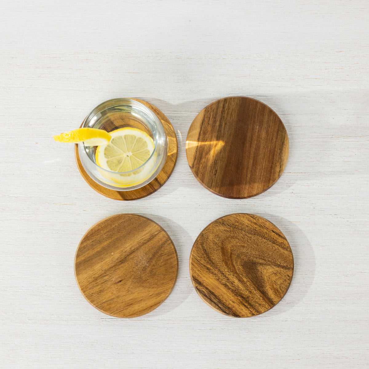 Gather 4 Piece Acacia Coaster Set shown from above with four round wooden coasters and a glass of lemon water resting on one coaster.
