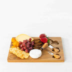 Gather 4 Piece Acacia Board & Knife Set featuring a natural acacia wood serving board styled with cheese, crackers, grapes, strawberries and dried fruit, with three coordinating cheese utensils.