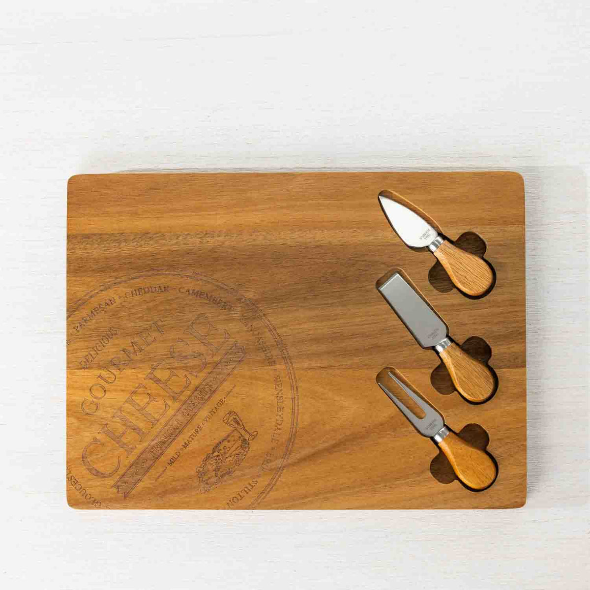 Gather 4 Piece Acacia Board & Knife Set featuring an engraved natural acacia wood serving board with three cheese utensils neatly stored in specially shaped recessed compartments.