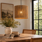 A cylindrical pendant light with a woven straw or jute shade hangs over a wooden dining table in a bright, modern room with a window, framed landscape print, and a vase of greenery.