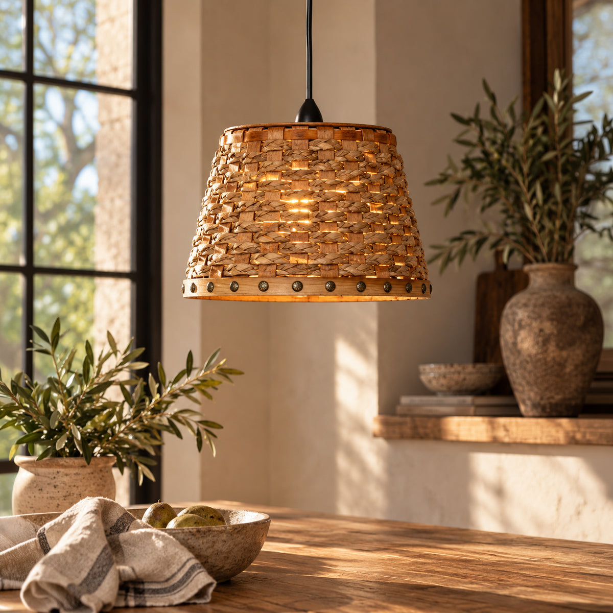 A woven natural-fiber pendant light shade hangs over a wooden table in a warm, sunlit room with plants and rustic decor.