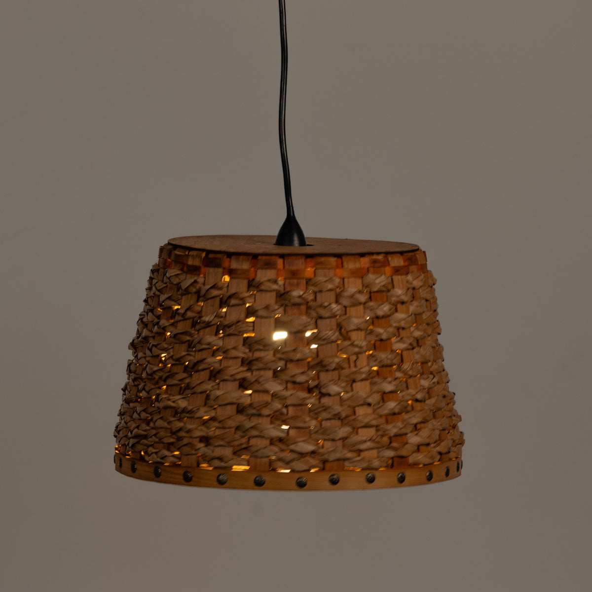 A woven natural fiber pendant lampshade with a black hanging cord, shown against a plain gray background with a warm light glowing through the textured weave.
