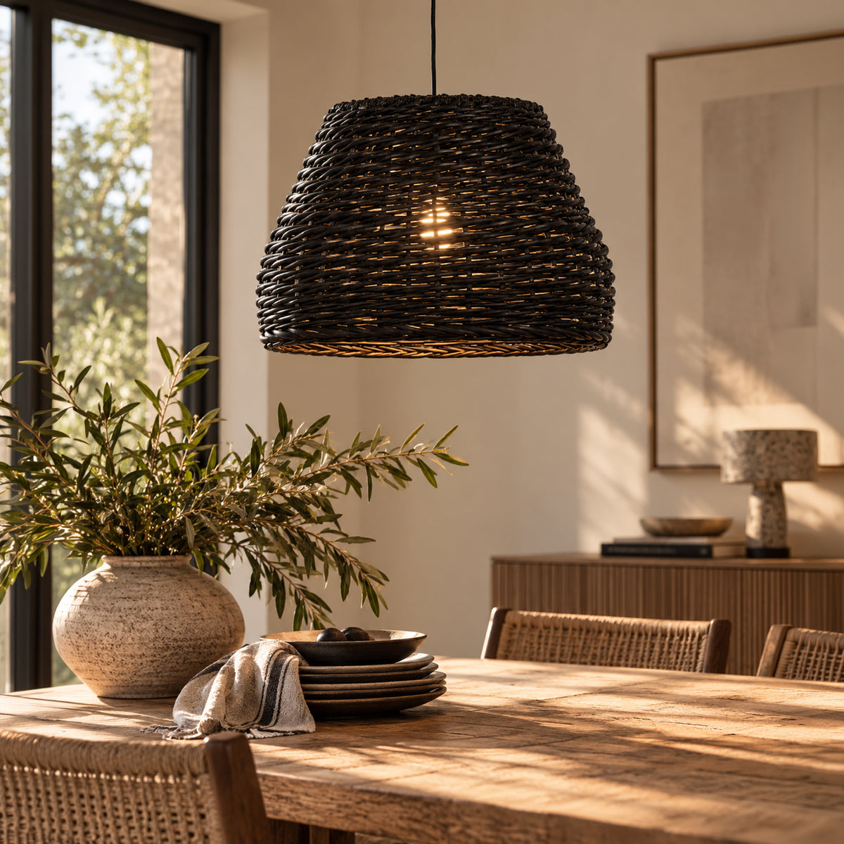 A dark woven wicker pendant light hangs over a wooden dining table in a warm, sunlit dining room with wicker chairs, a vase of greenery, and a sideboard in the background.