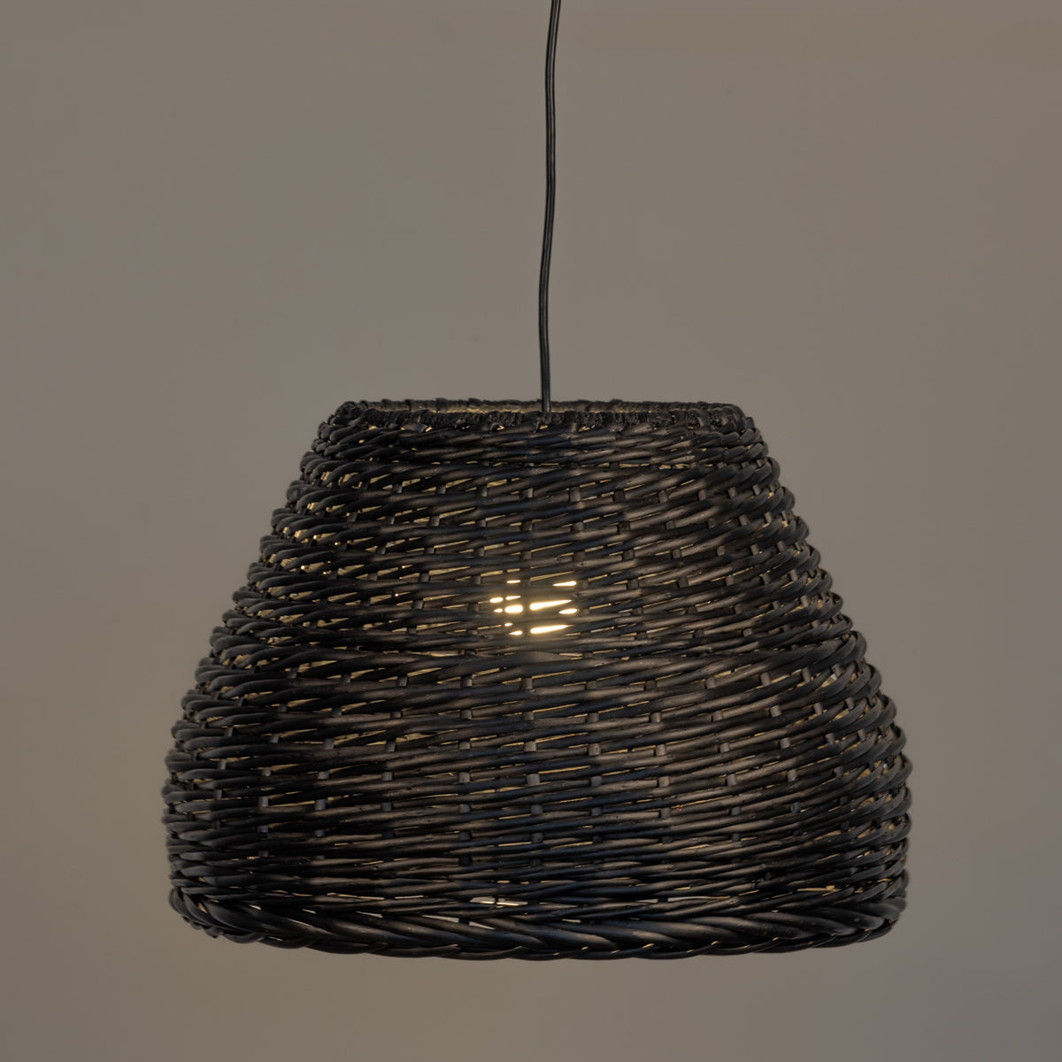 A dark woven wicker pendant light shade hanging from a black cord against a plain warm gray background.