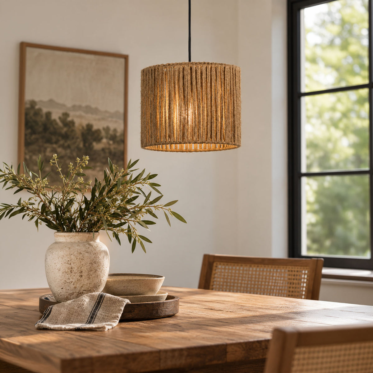 A warm, glowing cylindrical pendant light with a natural rope shade hangs over a wooden dining table in a bright, modern room with a window, chairs, and a vase of greenery.