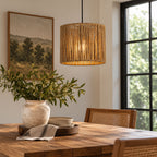 A warm, cylindrical pendant light with a woven natural fiber shade hangs over a wooden dining table in a styled room with a framed landscape print, a vase of greenery, and woven chairs.