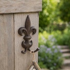 A decorative dark metal fleur-de-lis wall hook is mounted on a weathered wooden post, holding a hanging string ornament against a softly blurred garden backdrop.