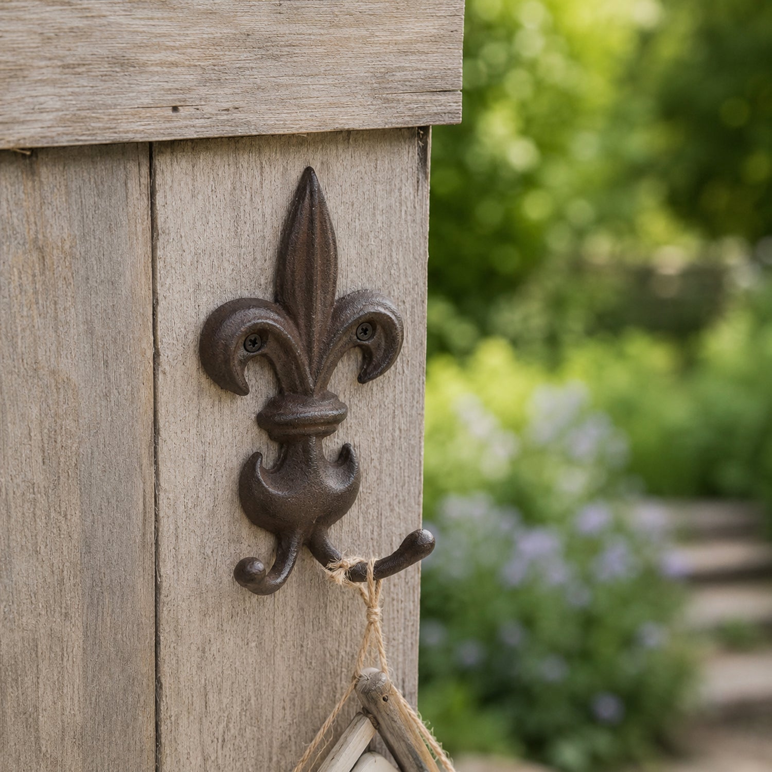 A decorative dark metal fleur-de-lis wall hook is mounted on a weathered wooden post, holding a hanging string ornament against a softly blurred garden backdrop.