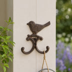 A decorative dark metal wall hook featuring a perched bird and two curved hooks is mounted on a light-colored post, with a hanging rope loop and a blurred garden background.