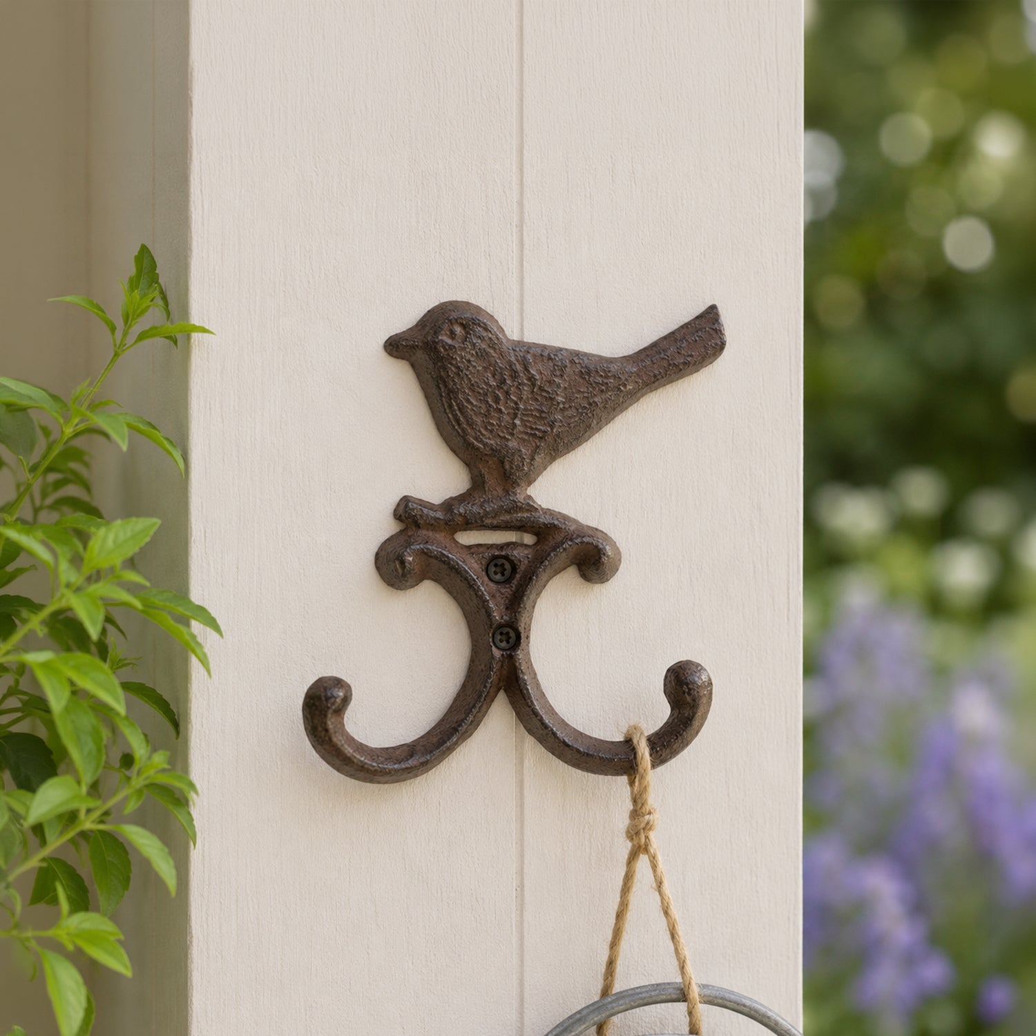 A decorative dark metal wall hook featuring a perched bird and two curved hooks is mounted on a light-colored post, with a hanging rope loop and a blurred garden background.