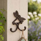 A decorative dark metal wall hook featuring a dragonfly above two curved hooks is mounted on a light-colored post, holding a hanging string ornament against a softly blurred garden backdrop.