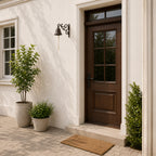A decorative dark metal bell with a rope pull hangs from an ornate scrollwork wall bracket mounted beside a wooden exterior door, with plants and soft sunlight completing the entryway scene.