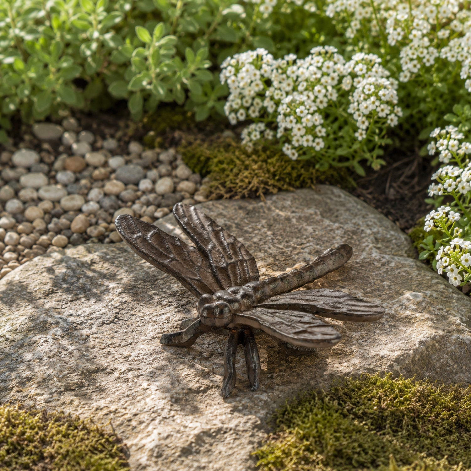 A decorative dark metal dragonfly ornament with textured wings rests on a large stone amid moss, pebbles, and small white flowers in a garden setting.