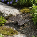 A decorative metal butterfly ornament with detailed patterned wings rests on the soil among rocks, moss, and garden plants in an outdoor garden setting.