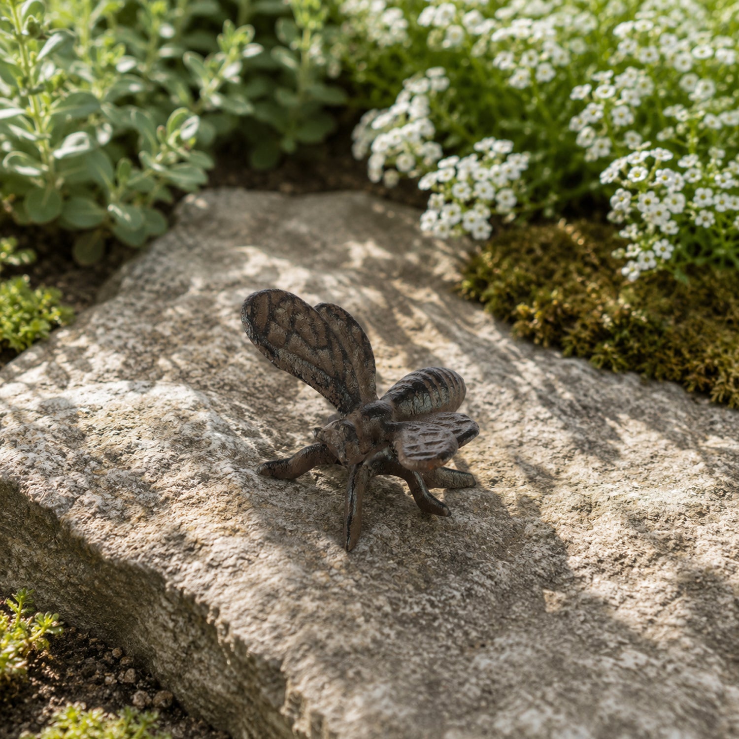 A decorative dark metal bee ornament with textured wings and legs rests on a large stone, surrounded by small flowering plants and greenery in a garden setting.