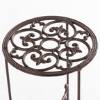 A close-up view of the top of an ornate dark metal plant stand shows an intricate scrollwork design with a circular frame and slender supporting legs against a plain light background.
