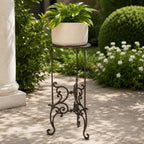A cream-coloured planter filled with lush green foliage sits atop an ornate dark metal plant stand on a sunlit garden patio surrounded by shrubs and flowering plants.