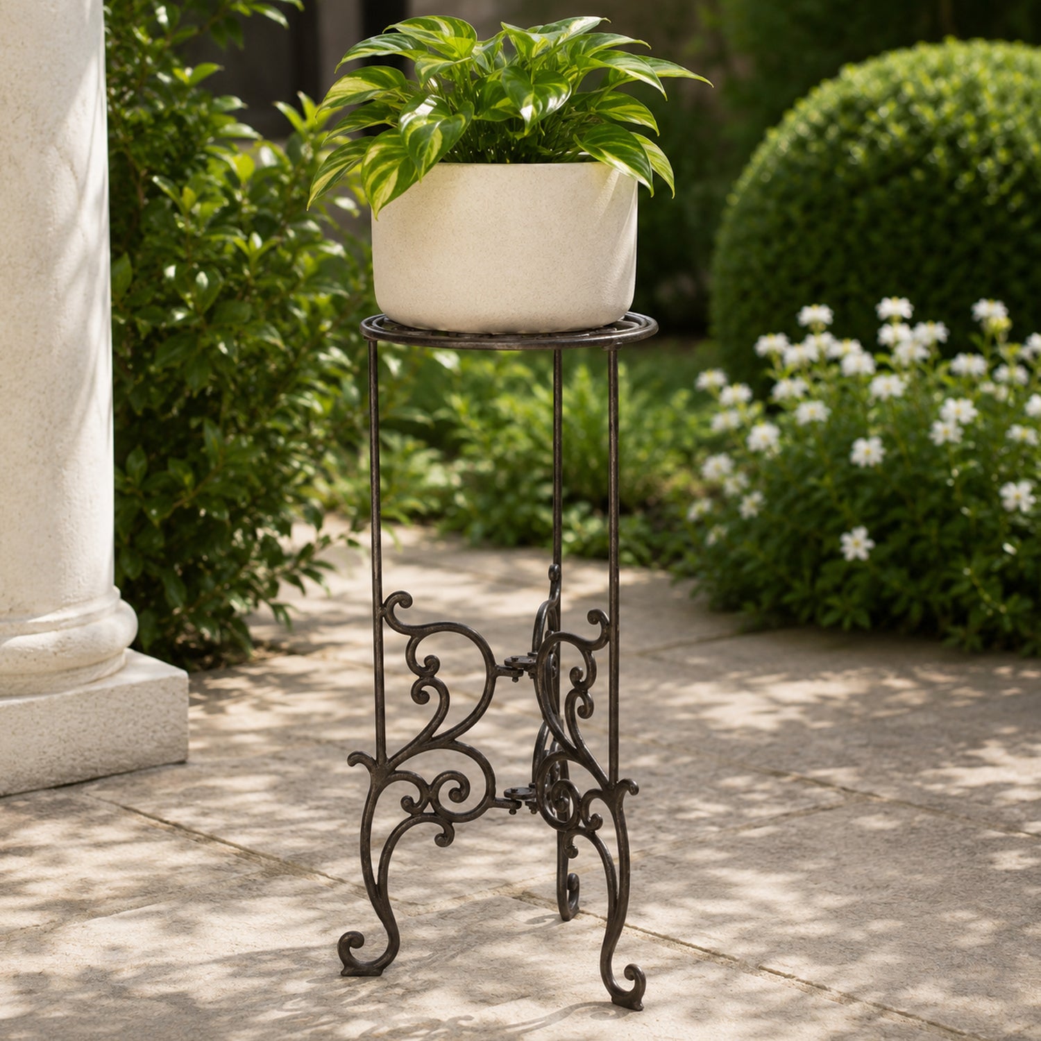 A cream-coloured planter filled with lush green foliage sits atop an ornate dark metal plant stand on a sunlit garden patio surrounded by shrubs and flowering plants.