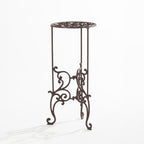 An ornate dark metal plant stand with a circular scrollwork top and decorative curved legs is shown against a plain light background.