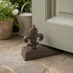 A decorative dark metal fleur-de-lis doorstop on a wedge-shaped base is positioned beneath a partially open door on a stone patio, with potted flowers and a watering can in the background.
