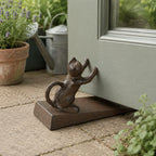 A decorative dark metal doorstop featuring a cat figure pushing against a partially open door sits on a stone patio, with potted plants and a watering can in the background.