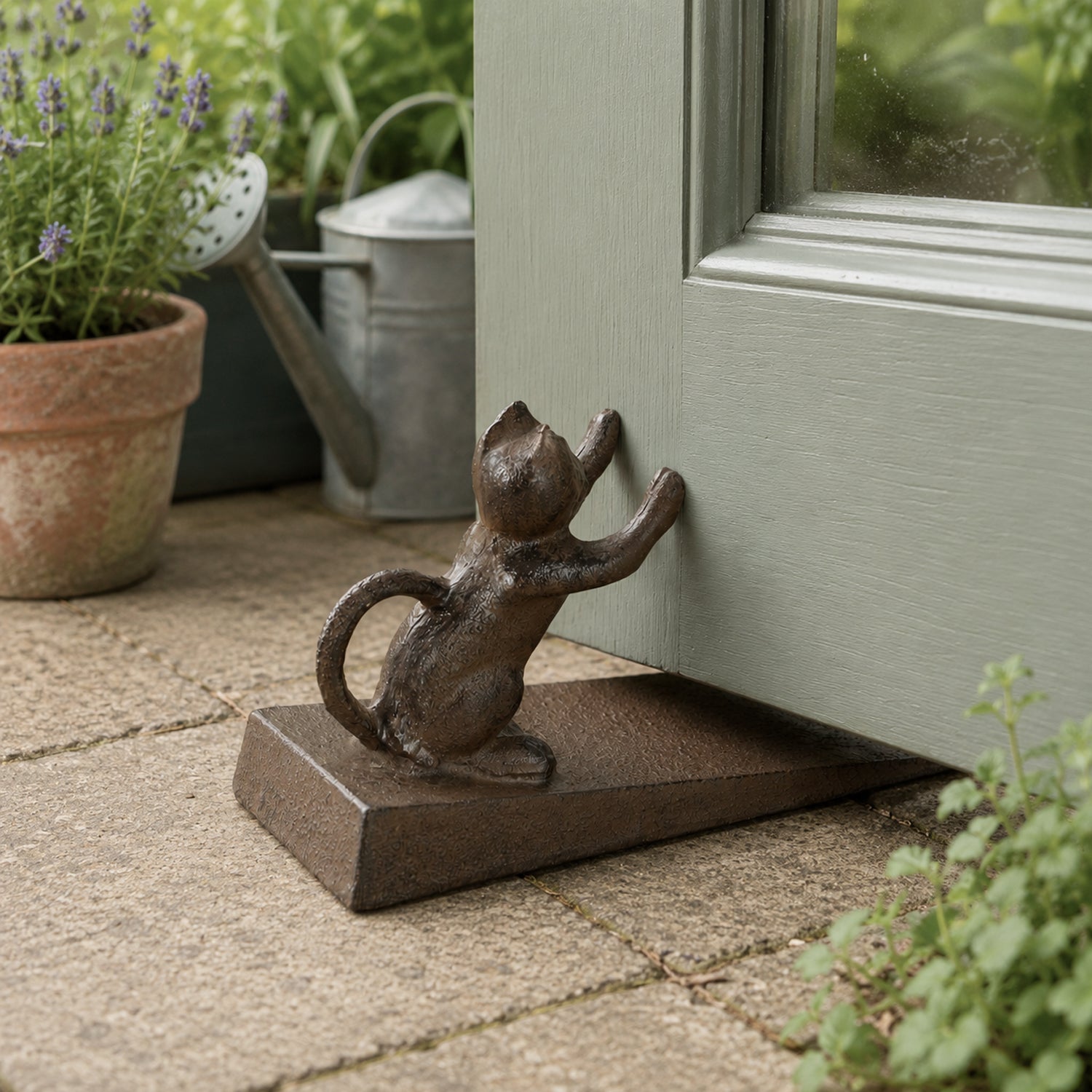 A decorative dark metal doorstop featuring a cat figure pushing against a partially open door sits on a stone patio, with potted plants and a watering can in the background.