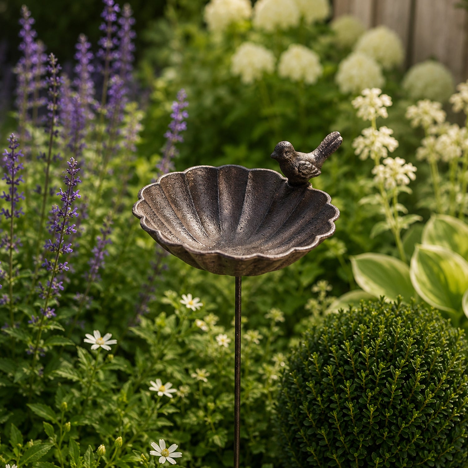 A close-up of a decorative metal garden stake with a perched bird accent is set among lush greenery and blooming flowers in a sunlit garden.