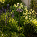 A decorative metal bird & flower garden stake with a small perched bird stands among lush flowering plants and greenery in a sunlit garden bed.