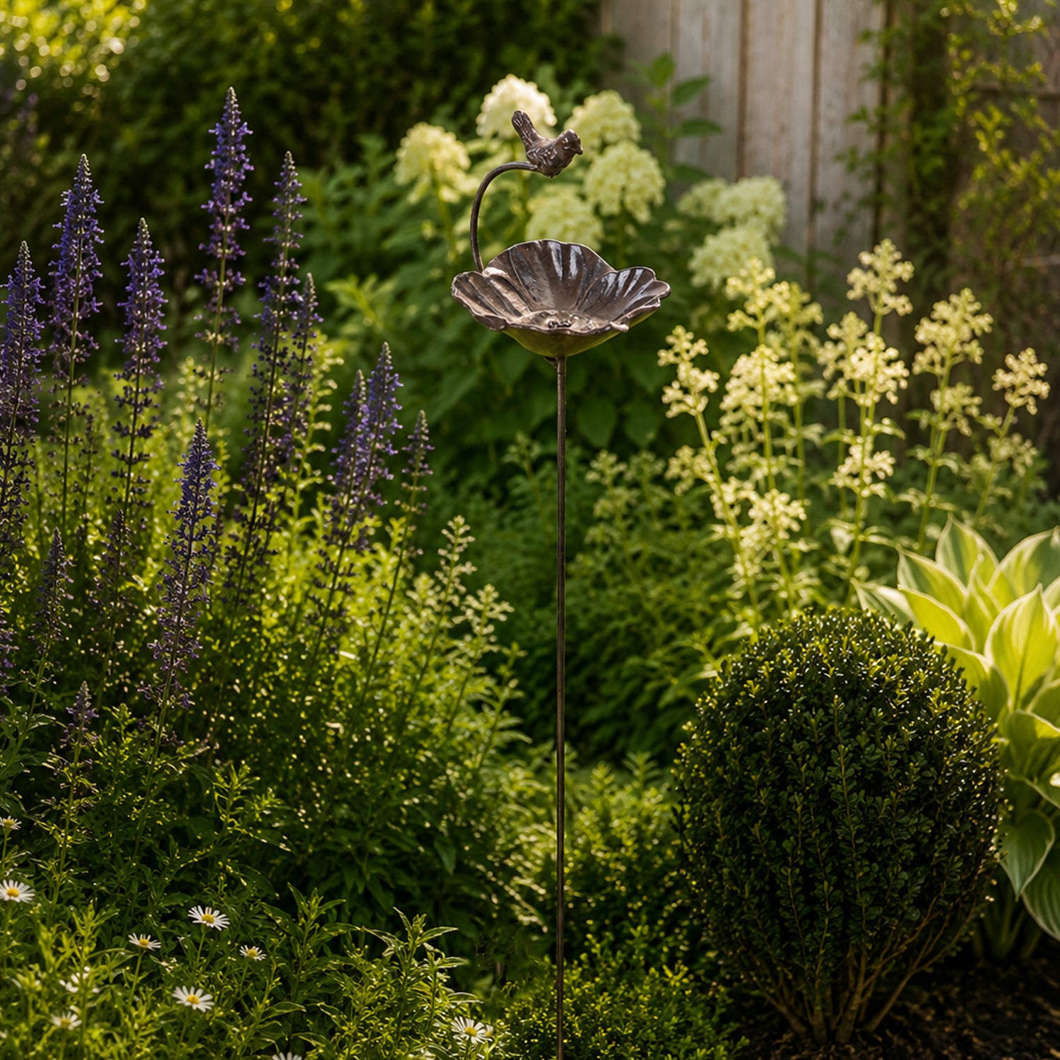 A decorative metal bird & flower garden stake with a small perched bird stands among lush flowering plants and greenery in a sunlit garden bed.