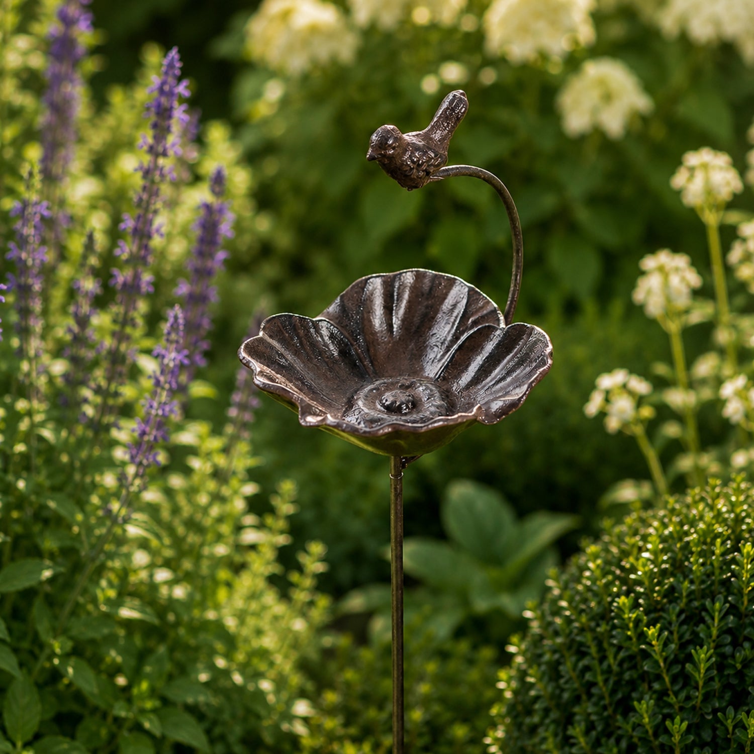 A decorative metal bird & flower garden stake with a small perched bird stands among lush flowering plants and greenery in a sunlit garden bed.