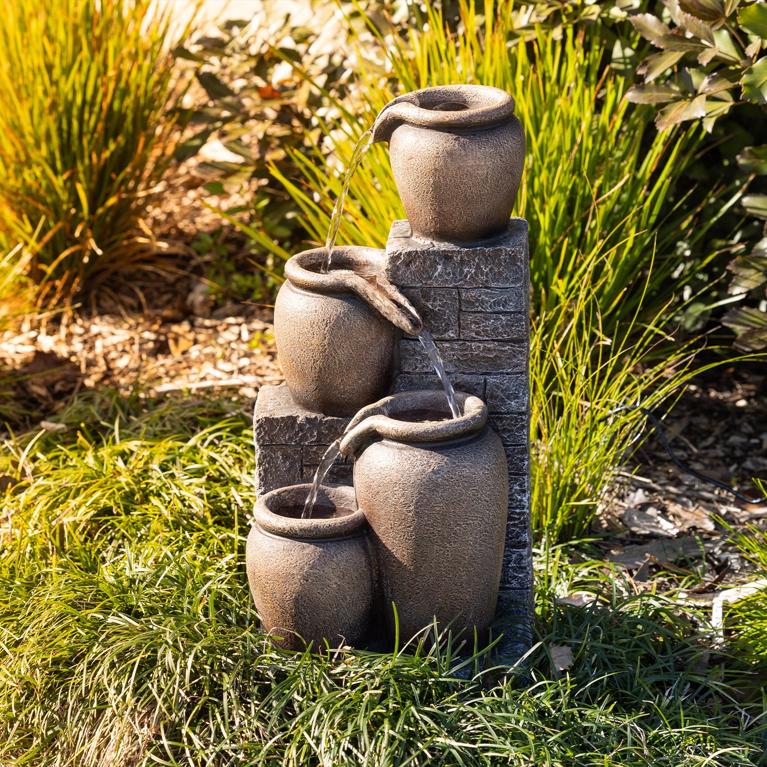 Outsidings Multiple Pot LED Fountain with cascading water flowing between textured stone-look pots, styled amongst lush garden greenery in a sunlit outdoor setting.