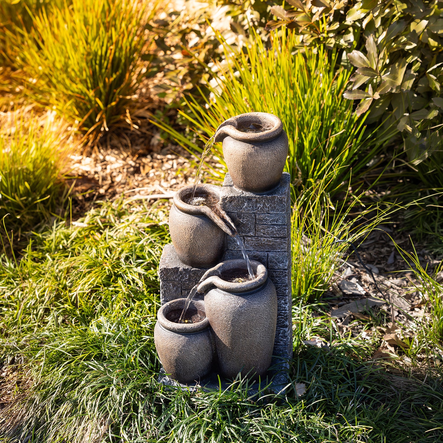 Outsidings Multiple Pot LED Fountain with illuminated cascading water displayed amongst ornamental grasses and garden plants in a landscaped outdoor setting.