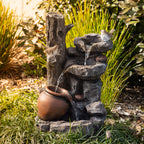Outsidings Bird & LED Fountain with cascading water flowing over natural rock formations into a rustic terracotta-style pot, accented by decorative birds in a landscaped garden setting.