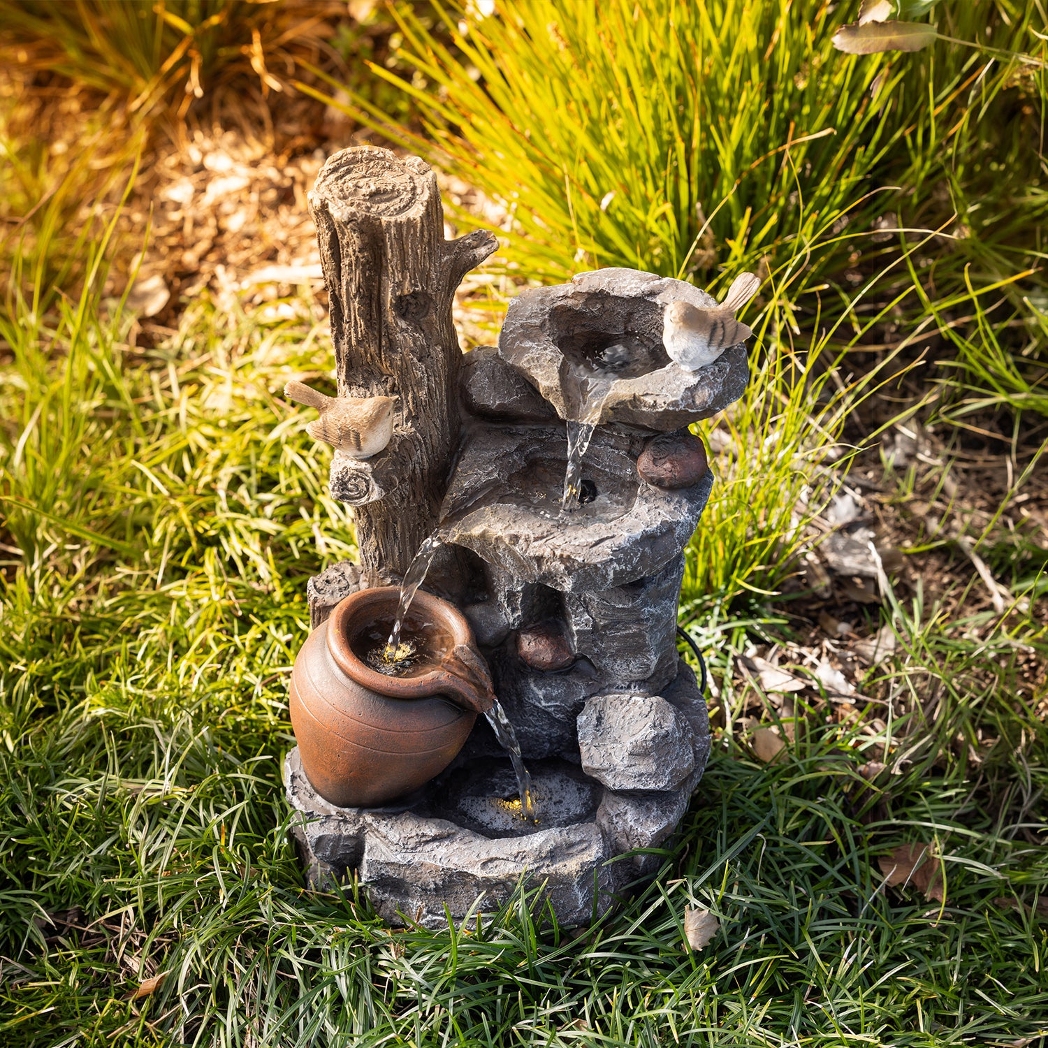 Outsidings Bird & LED Fountain with flowing water and warm LED illumination, nestled amongst ornamental grasses and garden foliage in a natural outdoor setting.