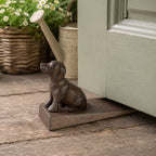 A decorative dark metal dog-shaped doorstop on a wedge-shaped base is positioned beneath a partially open door, with a watering can and flowering plants in the background.
