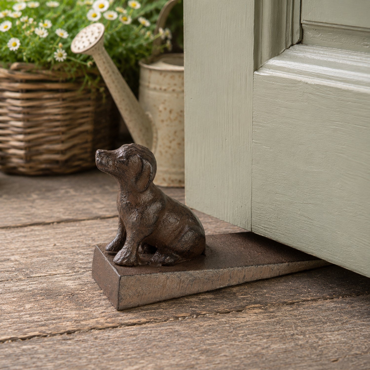 A decorative dark metal dog-shaped doorstop on a wedge-shaped base is positioned beneath a partially open door, with a watering can and flowering plants in the background.