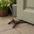 A dark metal doorstop in the shape of a simplified human figure is wedged beneath an open door on a stone patio, with garden accessories in the background.